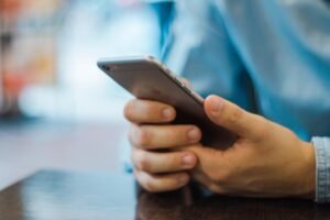 Homem de camisa jeans segurando o celular com as duas mãos, apoiado em uma mesa de madeira