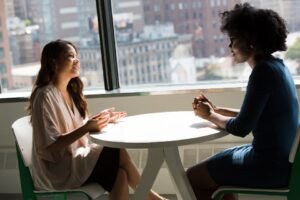 Duas mulheres conversando frente a frente em uma mesa redonda de escritório, com a cidade ao fundo pela janela