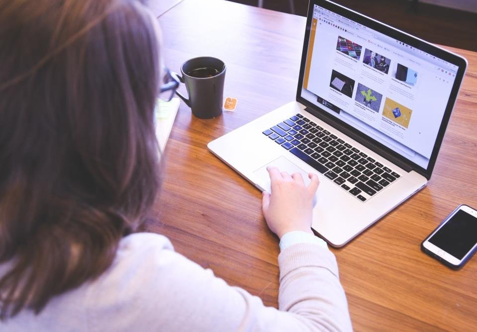 Mulher de costas trabalhando em um laptop sobre mesa de madeira, com xícara de café e celular ao lado